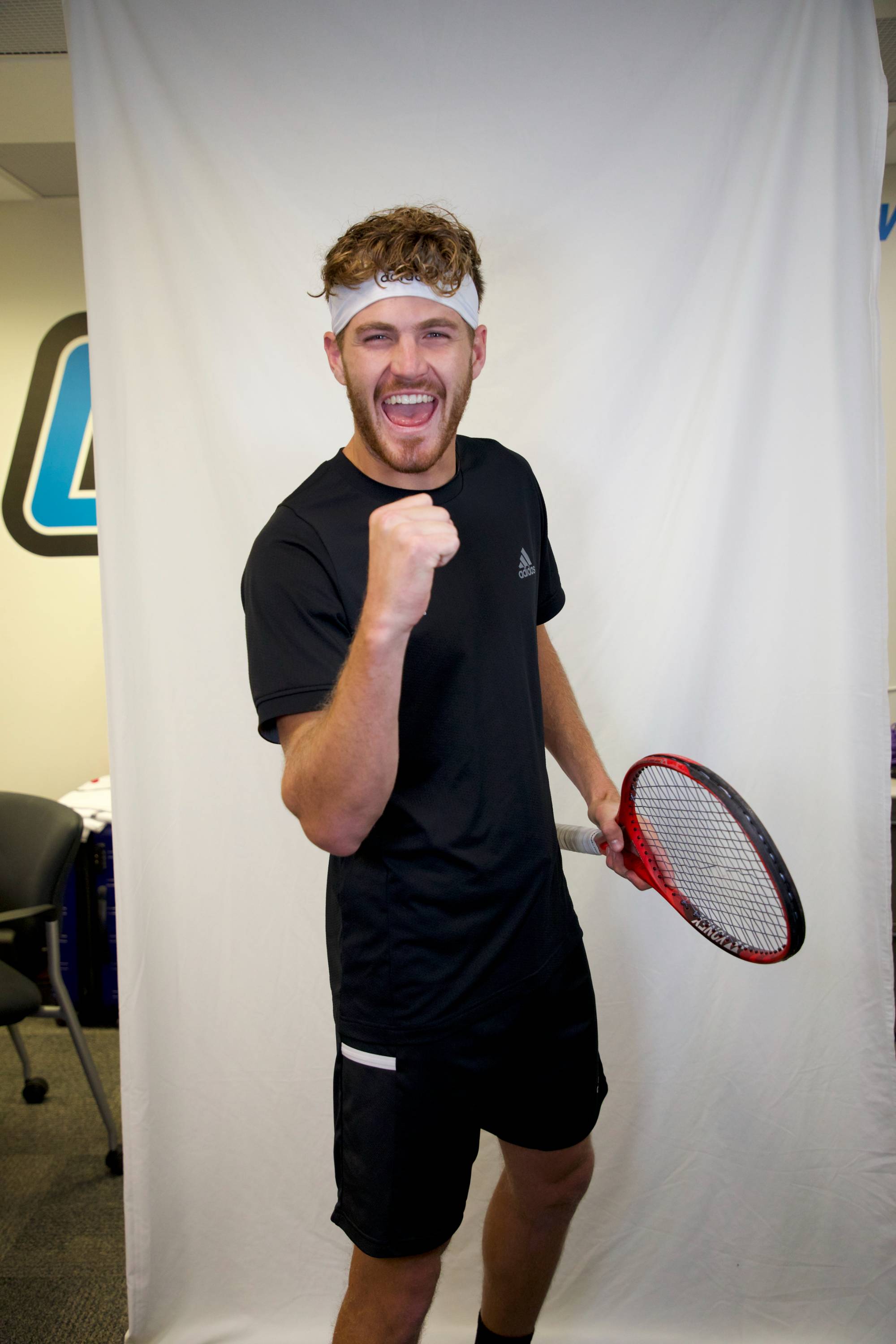 Men's Tennis Player Jack Dausman posing for a picture on media day, tennis racket in hand and fist cliched celebrating while smiling